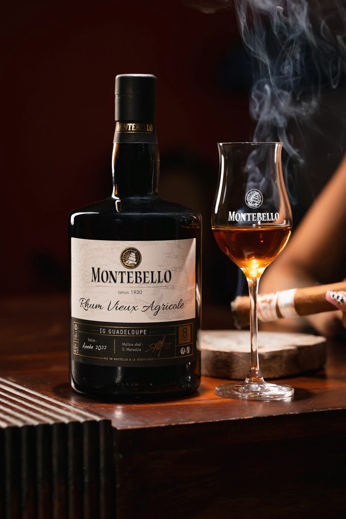 bouteille de rhum agricole de Guadeloupe de la distillerie Montebello, sur une tale à coté d’un verre a pied rempli de rhum. En fond, un cigare fumant