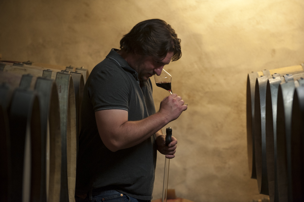 Pierre-André Frot goûte du vin dans un verre à pied dans une cave à vin, entouré de fûts en bois."