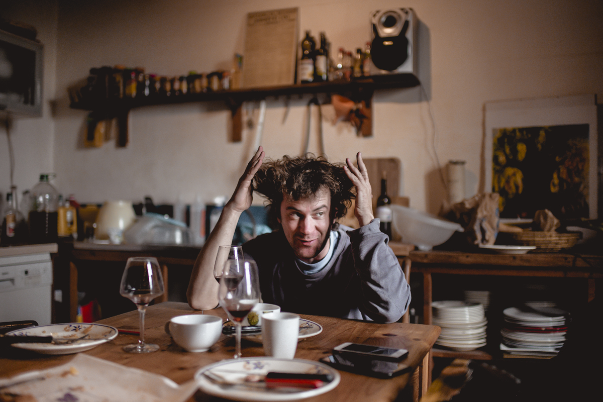 Julien Voogt ,les cheveux ébouriffés est assis à une table en bois dans une cuisine rustique, entouré de vaisselle et de verres."