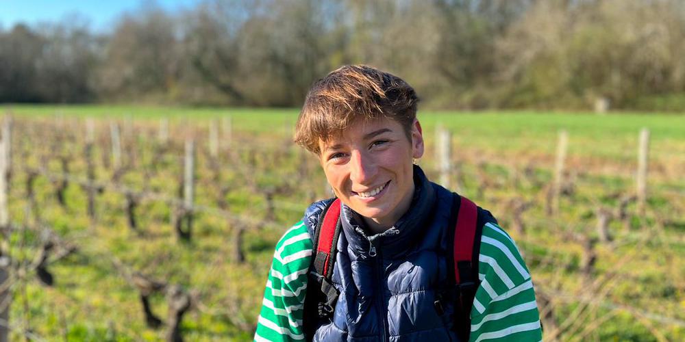 Lucie Mancais souriante en gilet bleu et chemise rayée verte se tient dans un vignoble, avec un paysage boisé en arrière-plan."