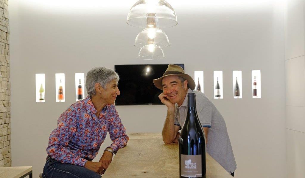 Deux personnes souriantes discutent autour d'une table en bois, avec une bouteille de vin au premier plan.